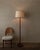 A calm, elegant vintage floor lamp with a slender teak-toned wooden stem, round base, and a clean fabric shade that gives...