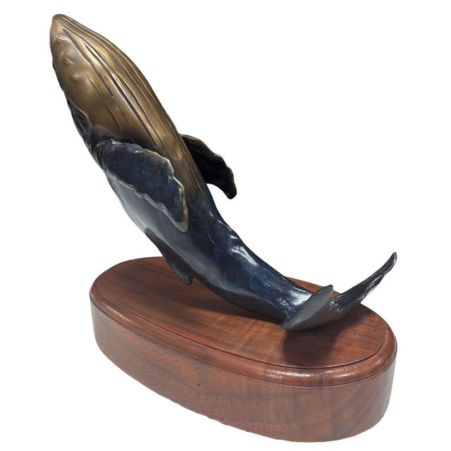 Elegant bronze whale sculpture mounted on a polished oak wooden base. The whale rises gracefully, its golden-toned back...