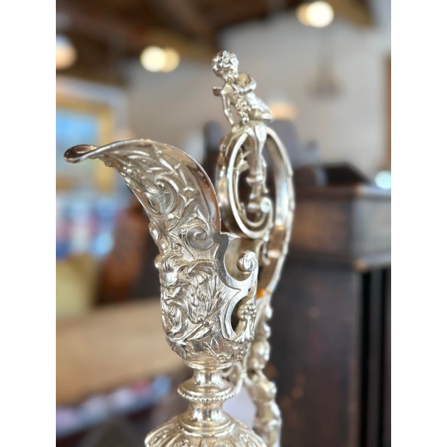 A monumental and exceptionally ornate silvered metal ewer with coordinating underplate, richly modeled throughout with...