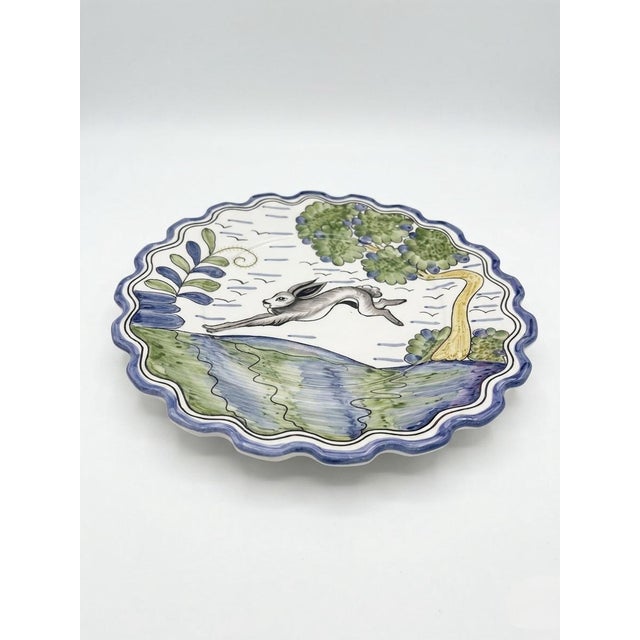This charming decorative plate features a lively, hand-painted scene of a leaping hare mid-stride across a rolling green...