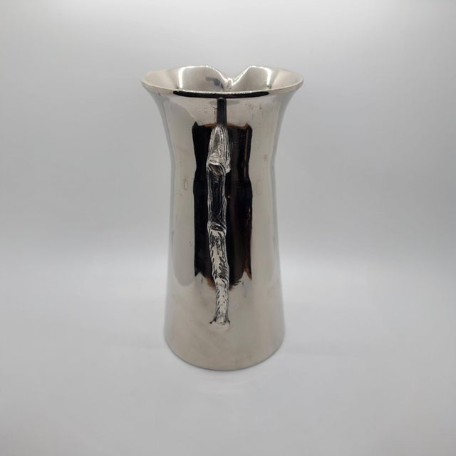 Polished silver tone pitcher features a unique twig-inspired handle with realistic bark texture. The sleek, tapered body...