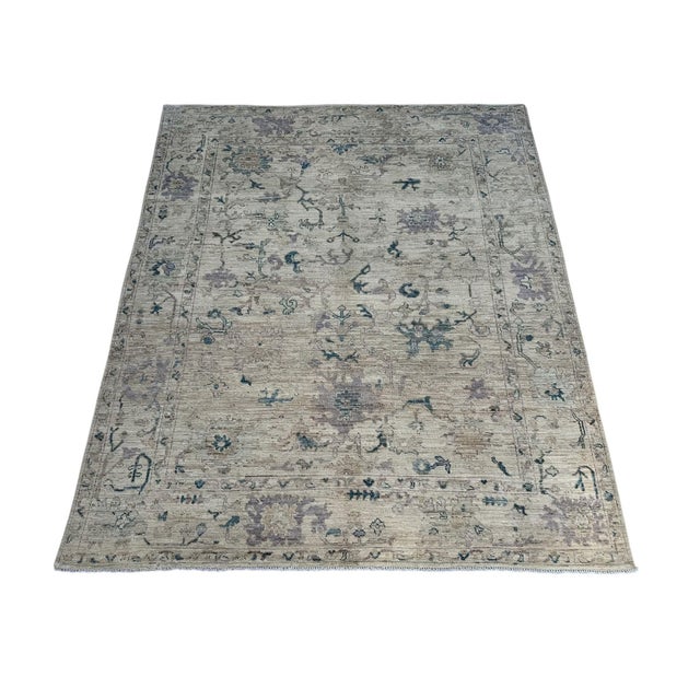 Muted lavender flowers and deep teal accents decorate the ivory field of this handmade Oushak wool rug. Fine taupe vines...