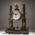 Small portico clock made of gray marble with gilded applications. The cylindrical movement is enclosed between two...