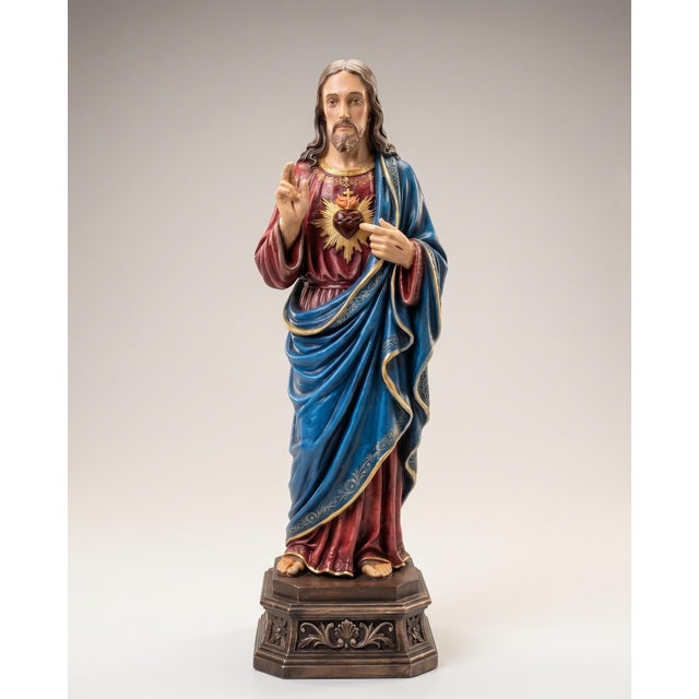 Jesus is depicted standing in a blessing gesture, with the Sacred Heart presented at his chest. A deep blue mantle, red...