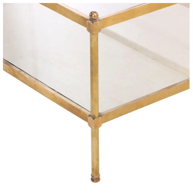 A coffee table featuring a slender brass frame with two clear glass shelves. The clean rectangular silhouette is accented...