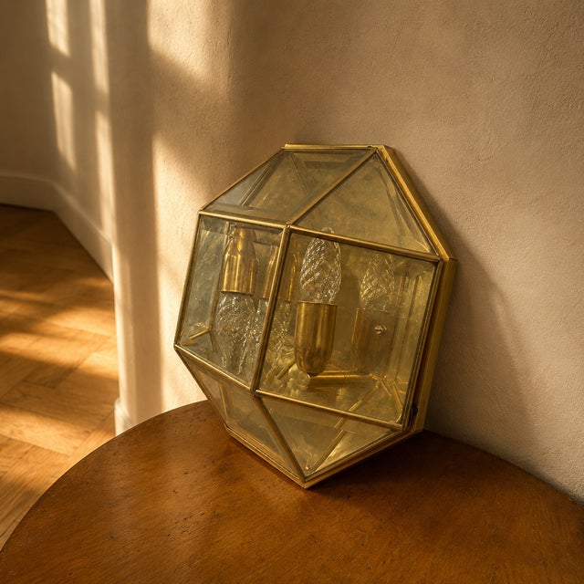 A vintage geometric wall light from the 1980s, featuring a faceted brass frame and clear glass panels. The multi-sided...