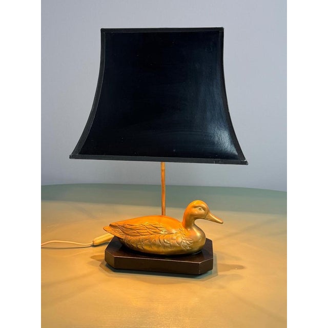 A striking vintage hollywood regency table lamp featuring a beautifully sculpted brass duck mounted on a substantial black...