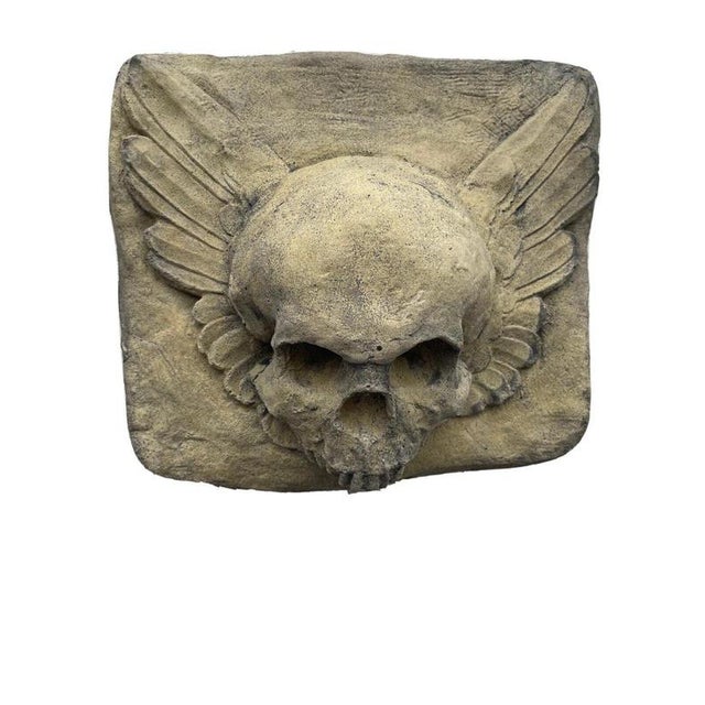 A striking Gothic-style wall plaque featuring a sculptural skull framed by dramatically outstretched wings. Richly...