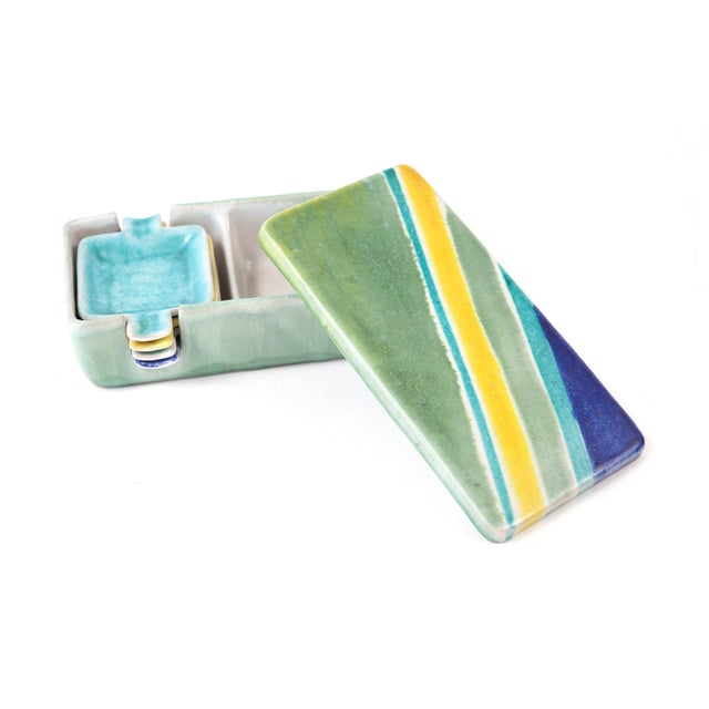 This celadon green ceramic box sporting vibrant stripes on its lid holds a set of four small individual cigarette ashtrays...