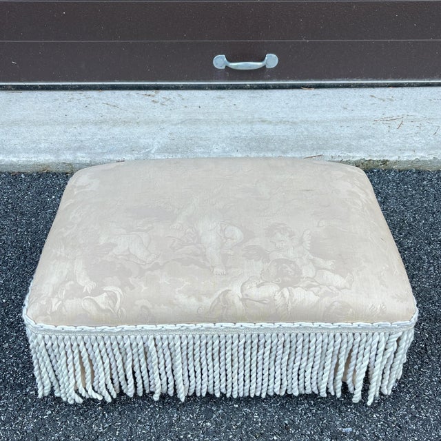A small mid 20th century Regency style ottoman or footstool having beige cherub patterned fabric with white tassels and...