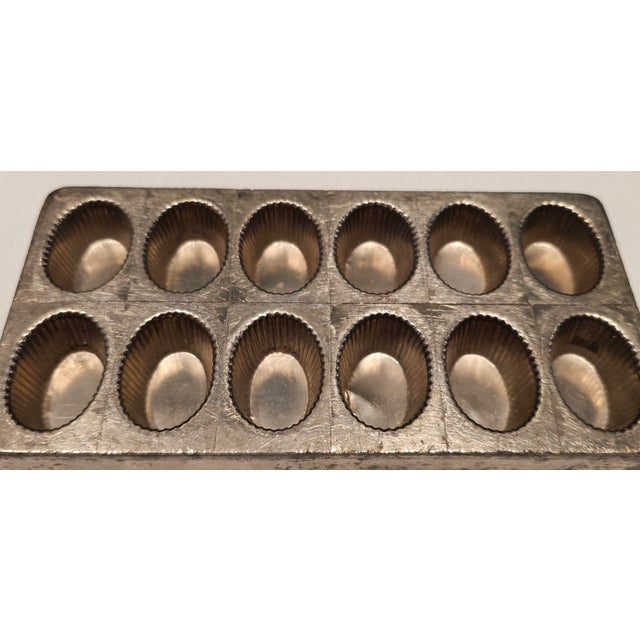 A beautifully aged antique French pastry mold featuring 12 oval, fluted cavities, stamped “NICKL / AQUI”, indicating...