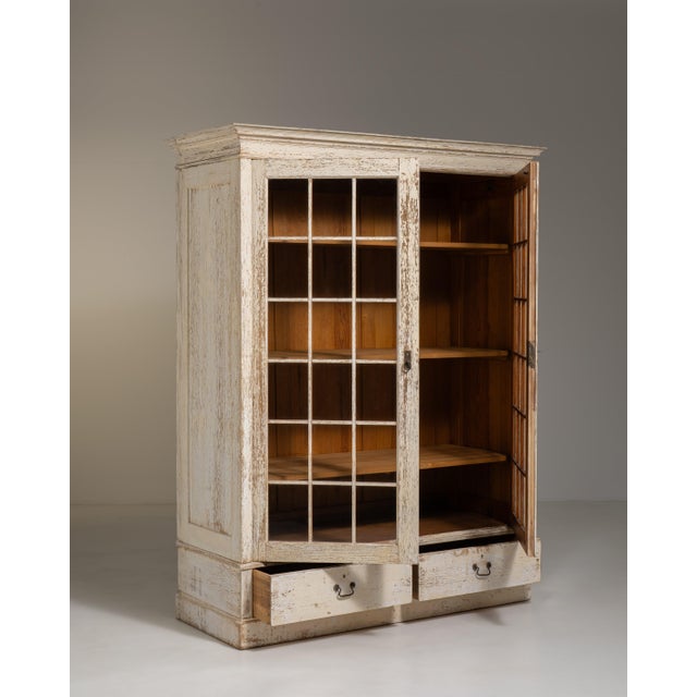 A tall vitrine featuring a softly aged white patina that enhances its decorative charm. The glazed doors allow for...