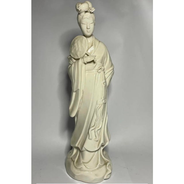 An elegant white porcelain figure of a lady holding a fan, distinguished by her softly lowered gaze, elaborate coiffure,...