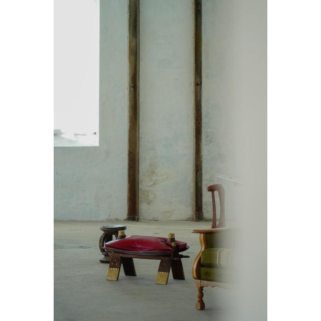 Distinctive camel saddle stool crafted in dark-stained wood with brass-mounted details and a rich red leather cushion....