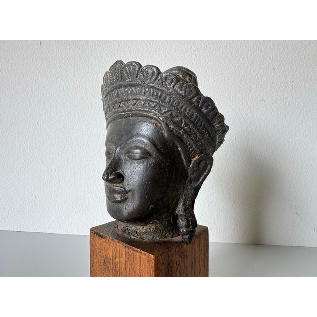 This 1970’s vintage cast bronze Buddha head sculpture is mounted on a simple wooden block stand, offering a serene and...