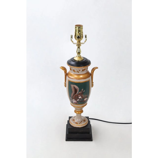 This vintage cream colored trophy cup lamp has leaf handles and a ring of foliage in gold color, with a small painting of...