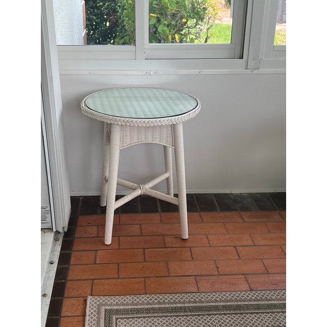 Vintage white wicker round side table equipped with a protective inset glass top surface. Table has top tightly braided...