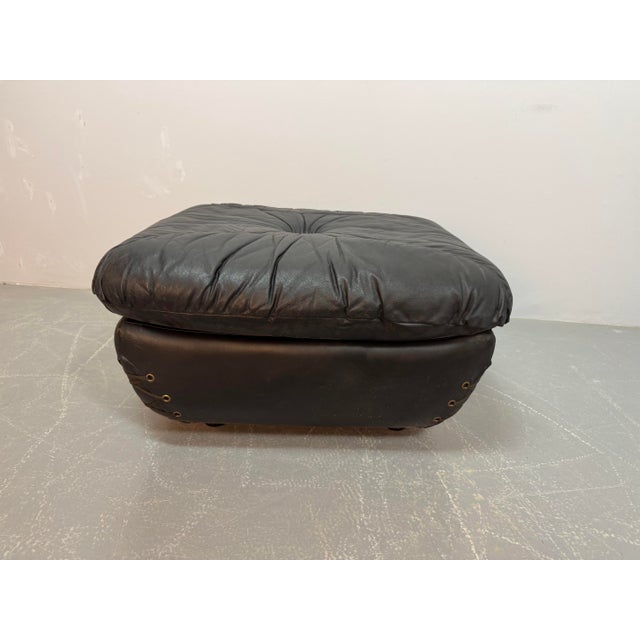 Mid-Century black leather pouf featuring distinctive cord lacing at the corners and decorative brass buttons on the...