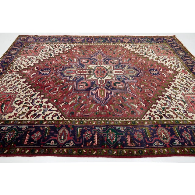 This Vintage Heriz wool rug has a red field, a purple-blue frame, and multicolor accents throughout the medallion motif....