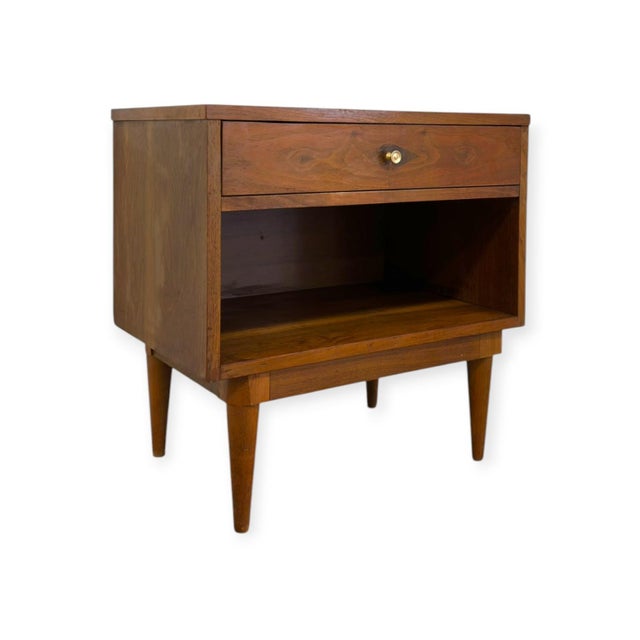 This mid-century modern nightstand features a rich warm wood grain finish, clean geometric lines, and iconic tapered peg...