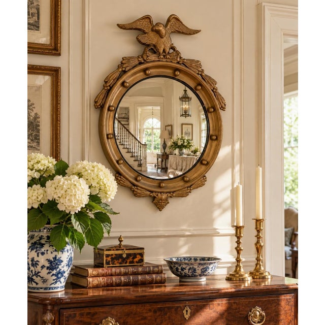 A Federal-style giltwood convex mirror crowned with a carved eagle, its wings outstretched above a laurel motif—an...