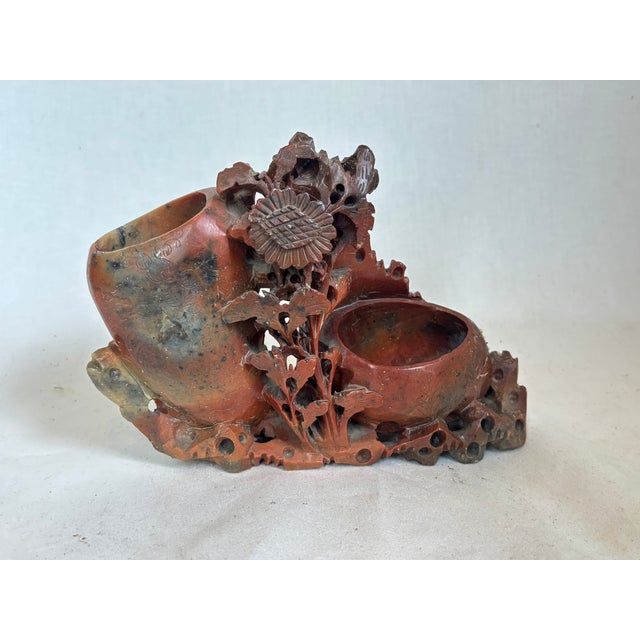 This carved soapstone vase depicts flowers has two holes. The vase is mainly brown with black coming though the glaze.