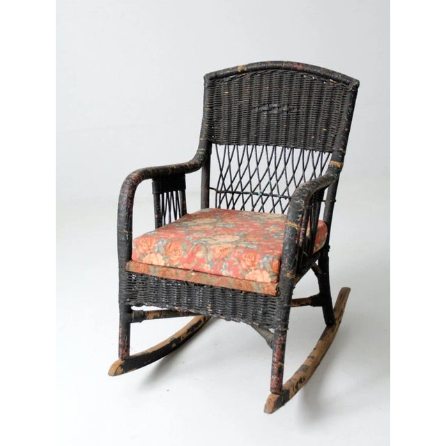 An antique wicker rocking chair featuring a sculptural armchair form and a distinctive black finish. The woven frame is...