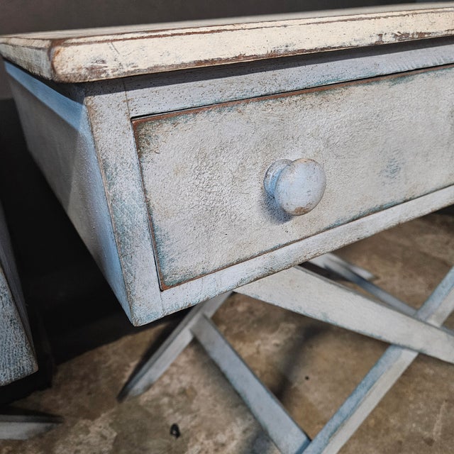 Pair of blue painted single drawer X frame side tables with blue accent line on top. The tables have a painted finish with...
