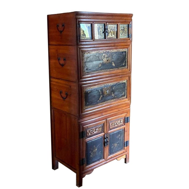 Tall Asian-style wooden cabinet featuring a warm reddish-brown finish with black lacquered panels, gold-tone decorative...