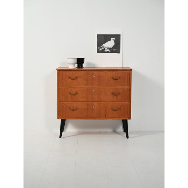 A 1950s teak chest of drawers with three drawers, double brass handles, and tapered black lacquered legs; a sober, graphic...