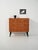 A 1950s teak chest of drawers with three drawers, double brass handles, and tapered black lacquered legs; a sober, graphic...