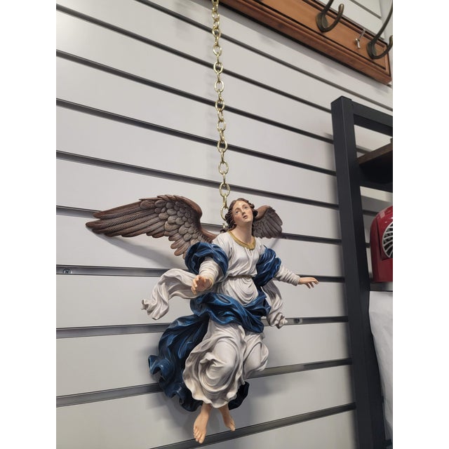 A winged angel is shown in a dynamic airborne pose, with open arms, softly modeled features and flowing blue and white...