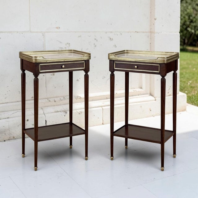 A pair of antique French Louis XVI end tables crafted of rich mahogany, each crowned with white marble set within a...