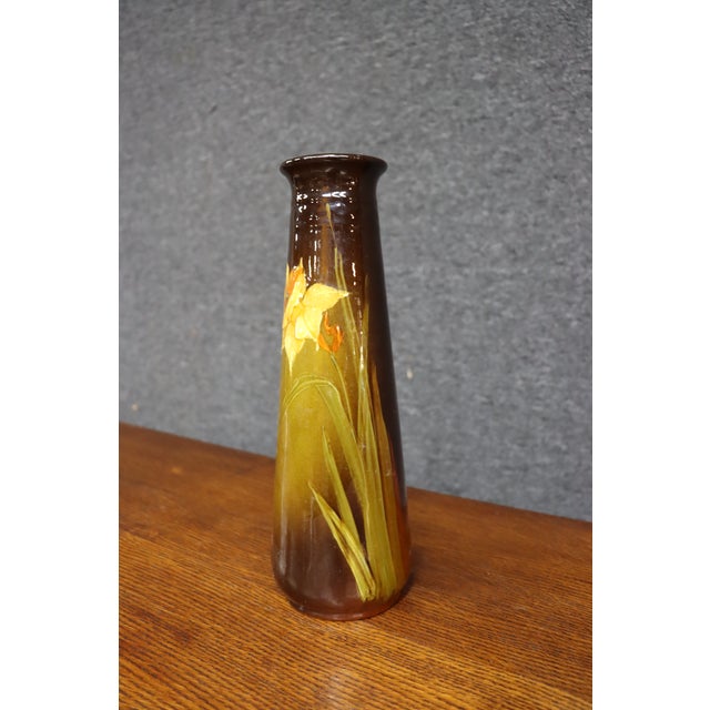 Weller Pottery vase featuring a tall, tapered form with a flared rim and glossy brown and golden ochre glaze. Decorated...