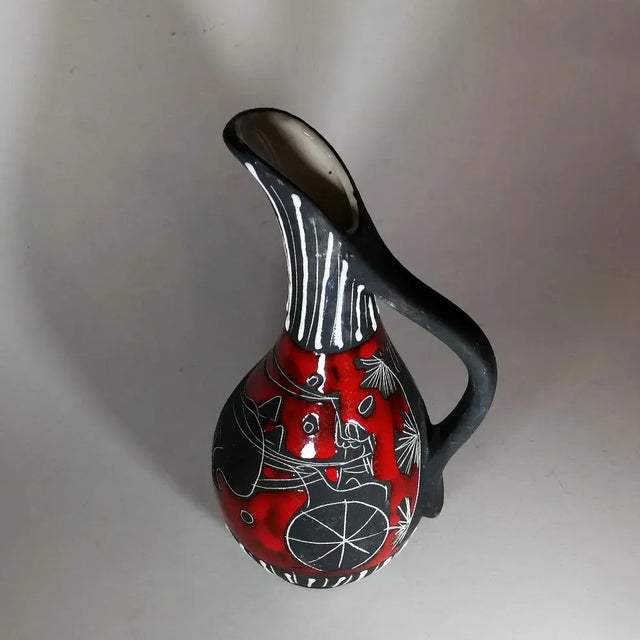 This carafe has a ceramic handle with scenes of life in red enamel and white on a black base. It has a label from Eturia...