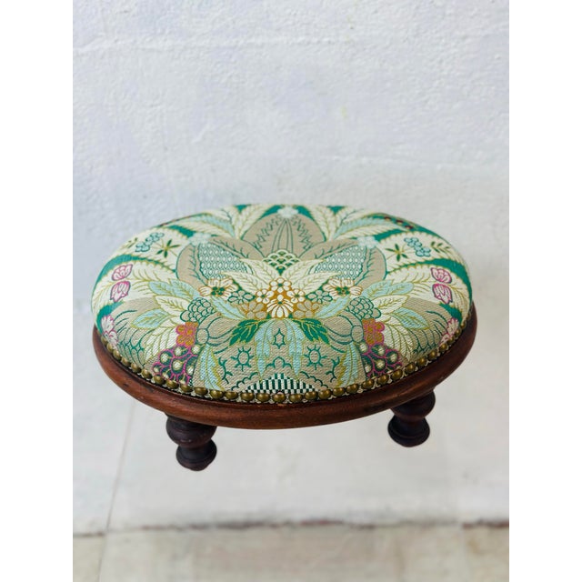 This is an antique round wooden footstool with turned legs and a padded upholstered seat. The cushion is covered in a...