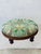This is an antique round wooden footstool with turned legs and a padded upholstered seat. The cushion is covered in a...