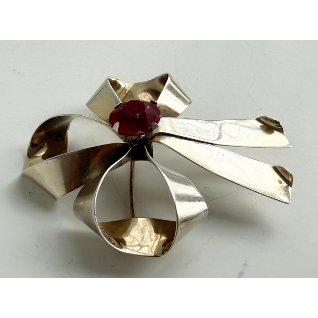 A darling sterling silver brooch, fashioned as a three-loop bow, set with a red, faceted gemstone.