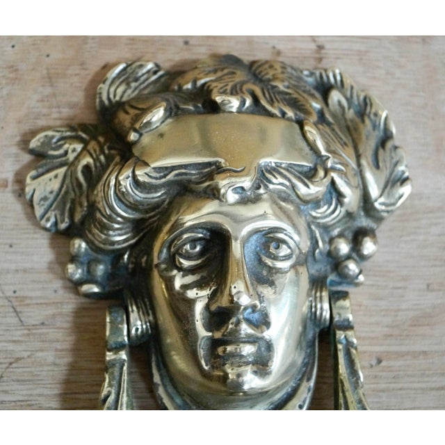 A cast brass door knocker shaped like the head of Dionysus crowned with an ivy wreath. Made from brass, remains in...