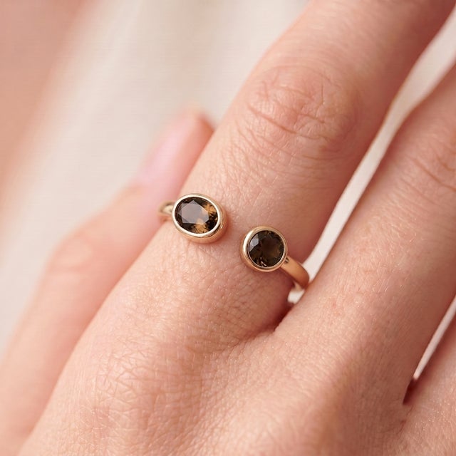 A bold, contemporary open-cuff ring featuring two earthy smoky-brown quartz stones — an oval-cut stone paired with a...