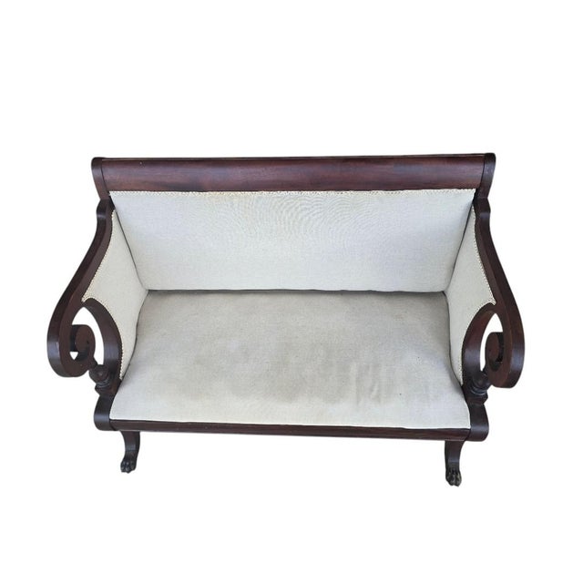 An elegant antique Empire Revival settee featuring dramatic rolled scroll arms, carved paw feet, and a richly figured...