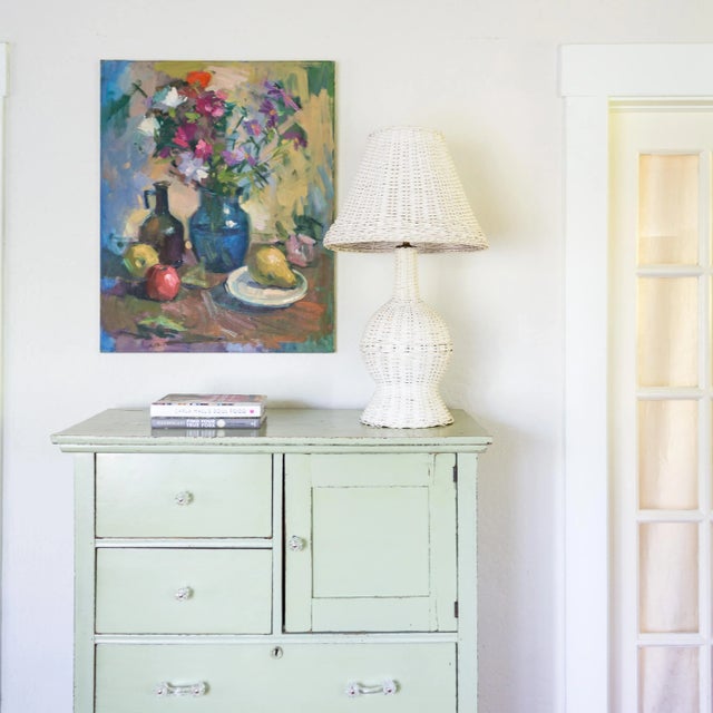 A large fruit and floral still life oil painting features a messy tablescape with a pear, apples, a glass jug, and a vase...