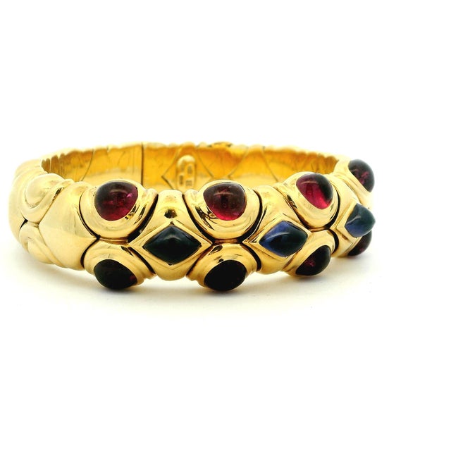 Substantial 18K gold bangle bracelet featuring a bold arrangement of cabochon rubies and sapphires set across a flexible,...
