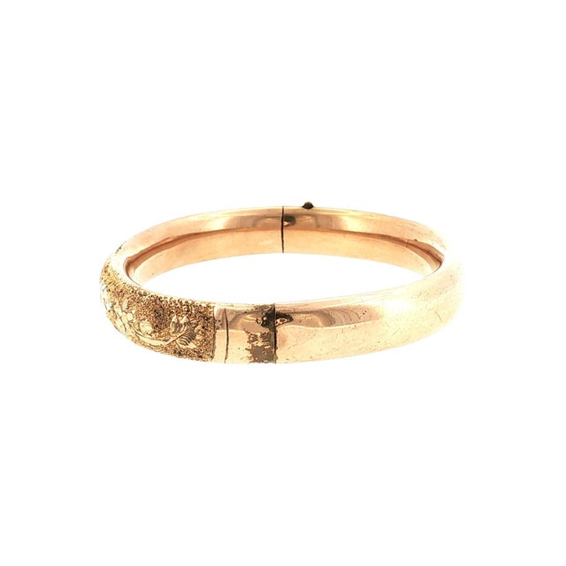 A late 19th-century 7.8mm wide gold-filled hinged bracelet. Features intricate floral engravings with a textured finish....