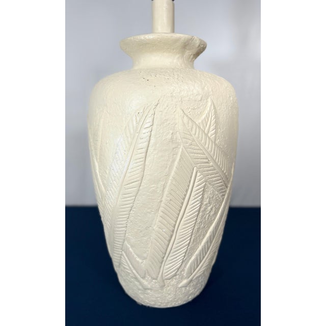 This striking table lamp features a heavy, urn-shaped body with a deeply recessed relief of overlapping palm fronds. The...