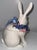 Fitz and Floyd ceramic white bunny is a lidded vessel. Pink and blue floral garland features a blue bow tied at the neck....