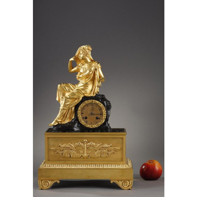 Clock in gilded and chiseled bronze representing a young woman sitting sideways dressed in the antique style, wearing a...