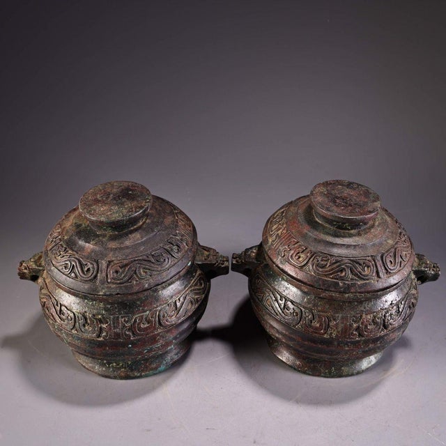This is a matching pair of cast bronze lidded vessels, featuring a rounded, bulbous body and a domed cover with a central...