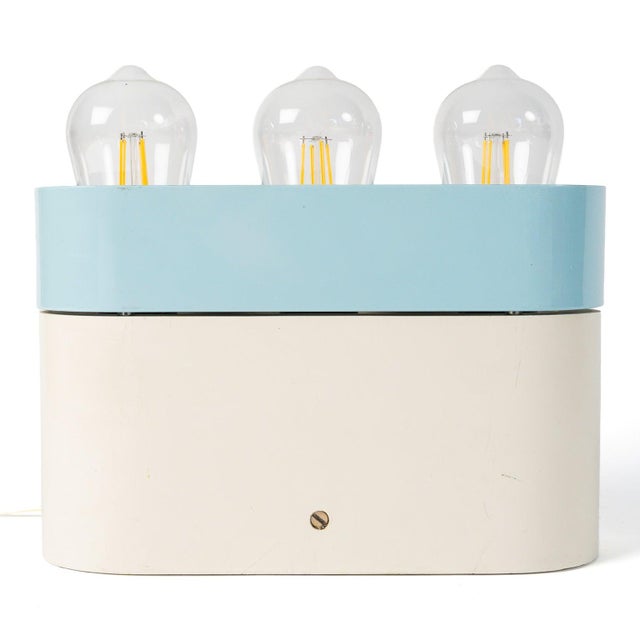 A horizontal, cream-lacquered wood and metal case, topped with an overhanging blue band that reveals three bare bulbs:...