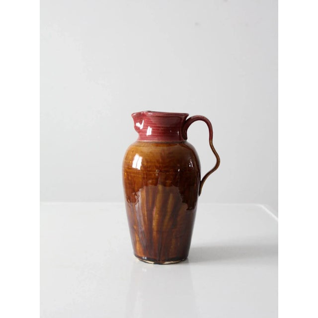 This hand-crafted studio pottery pitcher, dated 1983 and signed by the artist, features a warm brown and rose glaze with...